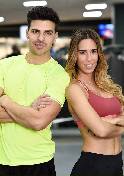 Een mannelijke en een vrouwelijke fitnesstrainer staan met hun armen over elkaar in de fitnessruimte en glimlachen